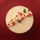 A round cream-colored cake with piped frosting and strawberry garnishes, photographed from directly above against a solid dark red background.