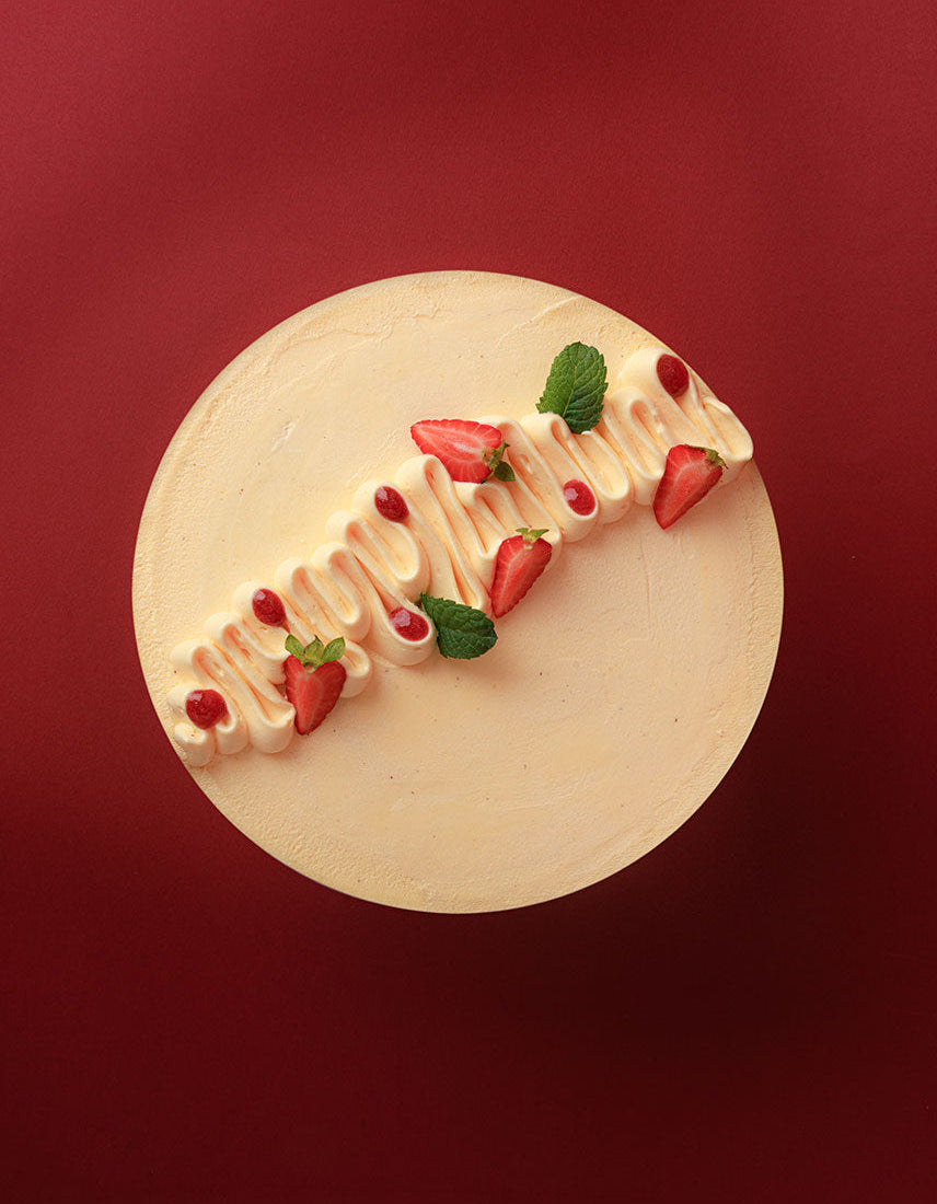 A round cream-colored cake with piped frosting and strawberry garnishes, photographed from directly above against a solid dark red background.